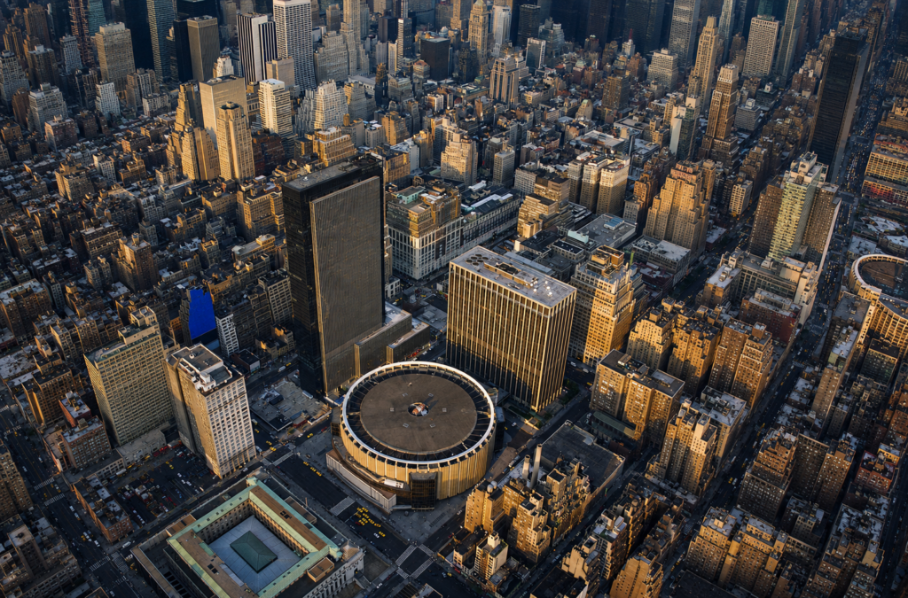 A wide aerial view of Midtown Manhattan at golden hour, featuring the circular roof of Madison Square Garden in the center of the frame. Surrounding the arena are densely packed skyscrapers and city blocks bathed in warm sunlight, with long shadows stretching across the streets below. The image captures the scale and density of New York City from above, with an expansive skyline extending into the distance.